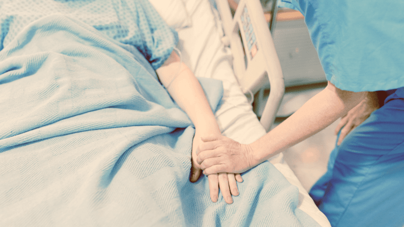 Person in scrubs holding bedridden patient's hand